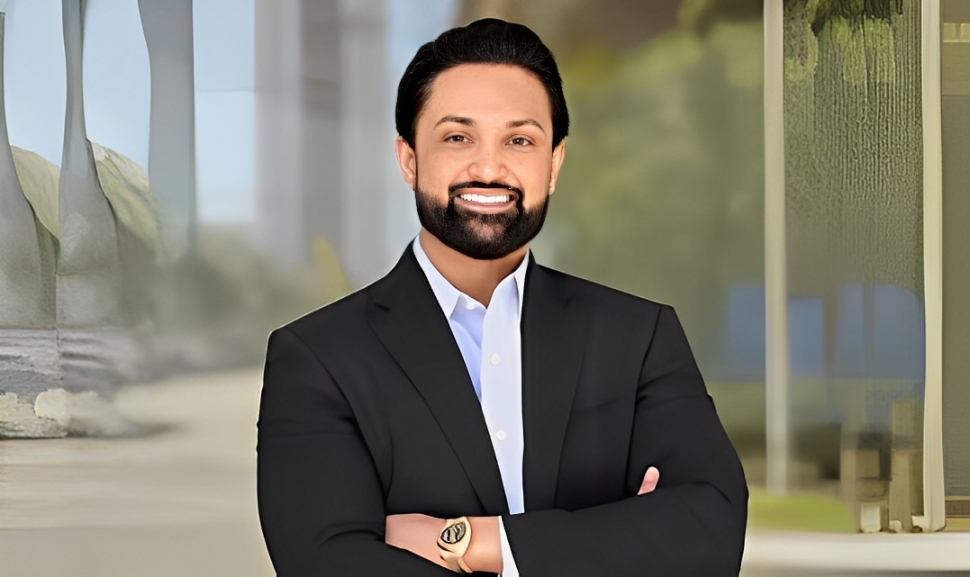 Bearded man in black suit smiling with arms crossed indoors.